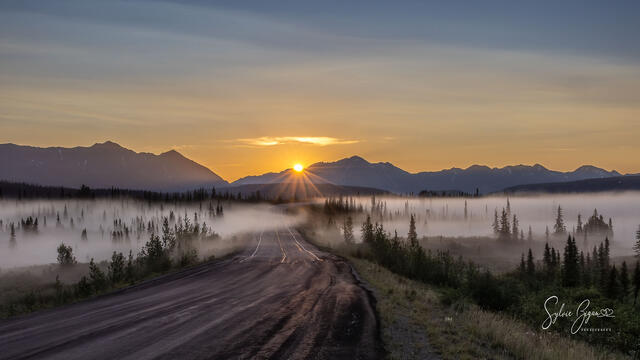 a road leads ahead to a foggy valley with the sun hiding behind a mountain range